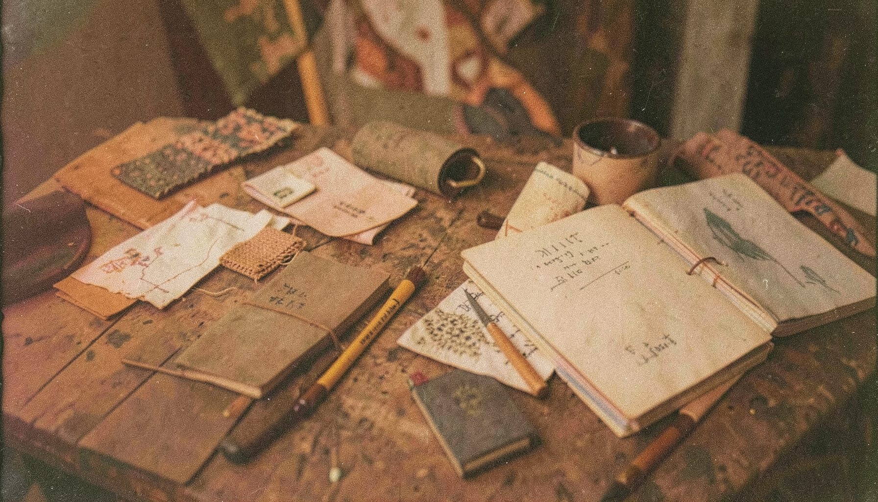A grainy film photo of handmade cultural artifacts and field notebooks arranged on a rustic table, earthy browns and muted ochres dominating the scene.