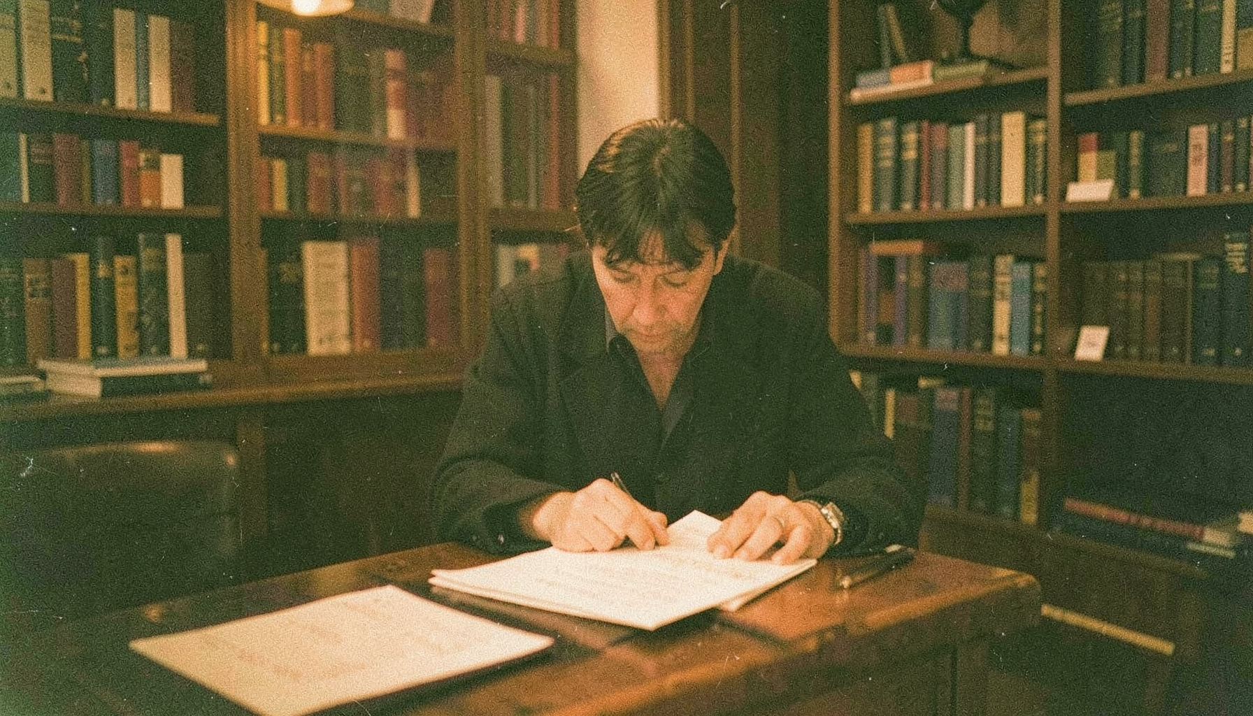 A grainy analog photograph of a person reviewing documents at a library table, warm amber light, scholarly atmosphere, deep greens and browns.