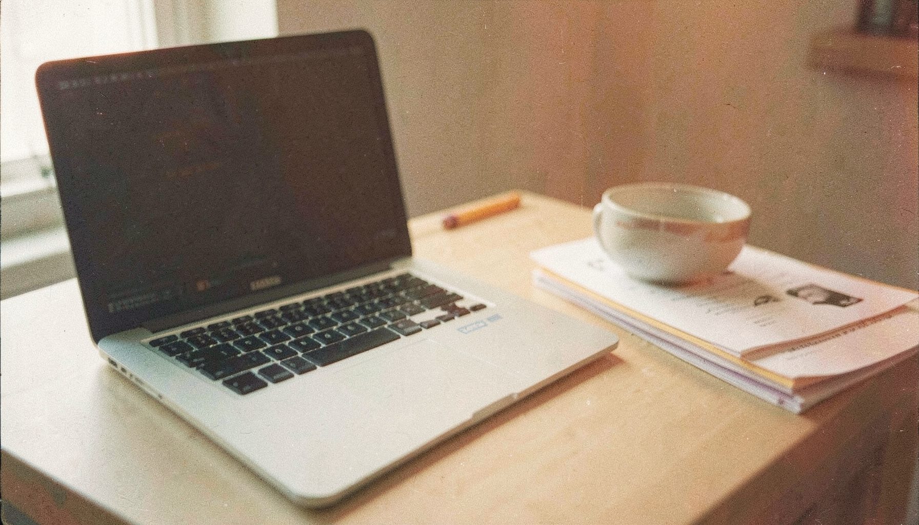A muted film photograph of an open laptop on a desk beside a coffee cup and stacked papers, soft morning light, grainy texture, slate and cream palette.